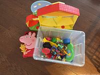Yellow and red plastic Peppa Pig house with open front. In front of it is a clear plastic bin containing various small toys.
