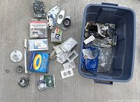 Photo of assorted plumbing and electrical items spread on the ground and in a blue storage bin showing various metal brackets, outlet covers, door knob, tape, wiring parts.