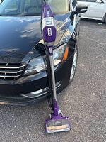 Full view of purple Shark Rocket vacuum cleaner standing upright against a black car, showing overall design and size.