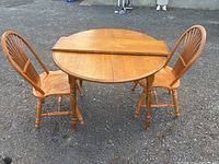 Round wooden dining table with two matching wooden chairs placed outdoors on gravel.
