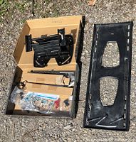 Box containing black TV wall mount bracket, metal support arms, and plastic bags with various mounting hardware components