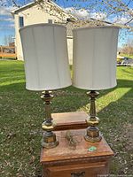 Two vintage metal base lamps with off-white fabric shades placed on wooden surface outdoors under daylight.