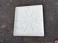 Top-down view of a square white plastic washing machine mat on the ground, showing ridges and some dirt.