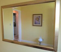 Full front view showing the entire large rectangular beveled mirror with gold gilt metal frame mounted on a yellow wall reflecting part of a room and framed artwork.