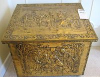 Front view of the firewood box showing brass embossed scene of people sitting around a table framed by trees.