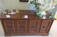Front angled view of the cedar chest illustrating the medium brown wood finish with four paneled doors, each fitted with antique brass ring pull handles, shown with decorative items on top.