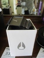 Black Nixon men's wristwatch on its white box with Nixon logo, showing square face and metal link band.