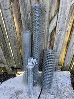 Four rolls of aluminum wire standing against a wooden fence on concrete. Rolls vary in height and mesh size, two rolls appear taller with finer mesh, two rolls shorter with larger meshes.