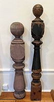 Two antique wooden balusters against a white wall and on wooden floor, one mauve, one black with worn paint showing natural wood beneath.
