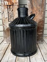 Front view of the antique black metal oil can with ridged body and shoulder, short angled spout, and attached lid with double hooks.