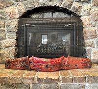 Front view of large antique wooden oxen yolk placed in front of a stone fireplace, showing its shape, red paint with wear and nail holes.