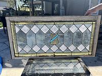 Full view of the antique rectangular stained glass window held outdoors showing the diamond pattern of textured glass and a central colored abstract motif in cream and blue, framed in white wooden frame with visible cracking and wear.