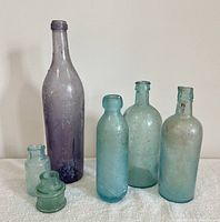 Group shot showing four antique tumbled glass bottles and two small glass inkwells arranged on a white surface against a plain wall. Bottles range in color from purple to aqua blue-green, varying in shape and neck style. Surfaces show weathering and patina indicating age.