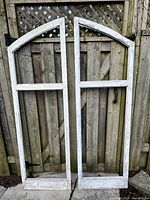 Two large white-painted wooden window frames leaning against a wooden fence, one has visible metal hinge attached at the bottom.