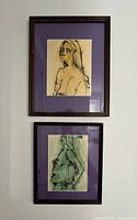 Two framed watercolour paintings of female figures with purple mats hung on a gray wall.