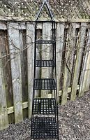 Tall black metal garden shelving trellis with five open lattice shelves shown against a wooden fence outside.