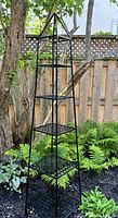 Full view of the tall black steel garden art piece with four lattice shelves and pyramid top, placed outdoors among garden plants.