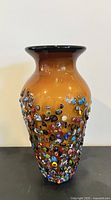 Front view of an amber-colored hand blown glass vase with bulging sides, decorated with colorful millefiori glass floral patterns scattered around the lower two-thirds.