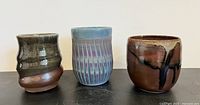 Group photo showing the three artist-made handleless mugs with distinct glazes: brown with black drips, blue with purple stripes, and brown-green textured.