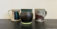Three artist-made mugs arranged side by side, showing variety in shape and design; middle mug is handleless and glazed dark brown to teal