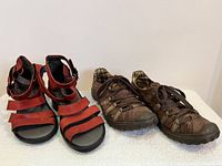 Side-by-side view of red Fly London sandals and vintage Rocket Dog shoes on a white surface against white wall background.