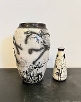 Front view of two handcrafted vases on a black surface against a white wall: one large raku-style pottery vase with black branch detailing and one smaller cream vase with black decorations.