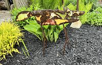 Full side view of steel garden fox sculpture in a garden bed with yellow and green plants and dark mulch showing the entire piece with visible legs and tail.