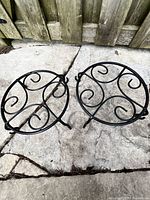 Two black metal planter bases with scrollwork, circular frame and three short legs, placed on stone ground.