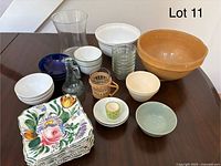 Photo showing a variety of kitchenware items including ceramic mixing bowls, floral square plates, glass carafe, small decorated egg cup, and clear glass tumblers arranged on a wooden table.