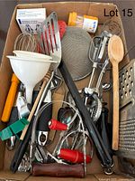 Top view of a box filled with various vintage and modern kitchen utensils such as graters, whisks, manual egg beater, wooden spoons, and peelers.