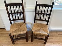 Pair of Spanish Colonial Revival dining chairs showing full front view with dark wood frames, double-row turned spindle backs with scalloped arches, and woven rush seats. Positioned side by side on wood floor with white baseboard behind.