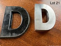 Top view of two metal sign letters 'D'; one black, 10 inches tall stamped by Adler Sign Letter Co., the other silver and 8 inches tall with brushed metal finish.