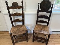 Pair of two vintage ladderback chairs side by side showing carved wood detail and woven rush seats, placed indoors against white wall.
