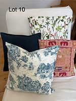 Photo shows four square throw cushions placed on a white chair or sofa. Visible cushions include botanical print with green and pink floral design, pink elephant motif cushion, blue toile pastoral scene cushion, and solid navy knit cushion.