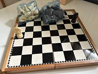 Open wooden folding chessboard showing glossy black and white checkered magnetic playing surface framed by decorative border. Two Staunton-style chess pieces placed on board and bags of pieces off to side.