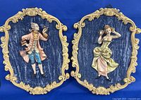 Front view of both velvet wall plaques with 18th century styled male and female figurines in pastel colors, mounted on gold decorative frames.