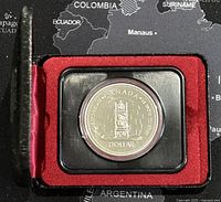 Commemorative coin displayed in protective capsule within black and red display case (lid open) against a map background.