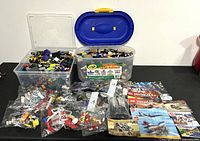 Photo showing two large plastic tubs filled with assorted LEGO bricks and several sealed numbered bags of LEGO parts, plus multiple LEGO instruction booklets.