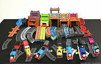 Lot displayed with six toy trains in a row, various curved and straight plastic tracks, and multiple colorful plastic buildings for train playsets.