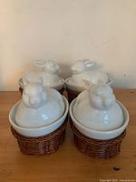 Four white ceramic bunny rabbit ramekins with detailed rabbit lids sitting inside brown wicker baskets on a wooden surface.