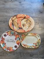 Photo shows three fall-themed platters on a wood floor: one round melamine platter with colorful leaves and 'Give Thanks' text, one oval melamine platter with leaf pattern, and one rectangular china platter with autumn leaf and floral border.