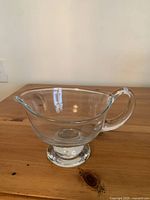 Photo of a clear glass gravy boat with a handle and pedestal foot on a wooden surface against a plain wall.