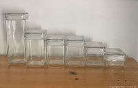Full set of six clear square glass apothecary canisters arranged in order of height from tallest to shortest on wooden surface.