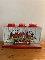 Front view showing the LANG flag basket artwork on the wooden votive box with three votive candle holders on top, each containing a red votive candle.