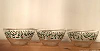 Three clear glass bowls with Christmas holly and red berry design arranged side by side on wooden surface.