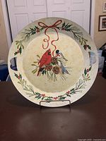 Front view of round metal serving tray showing handpainted winter scene with cardinal, bluejay, pinecones, holly, and red bow. Tray is displayed upright on stand.