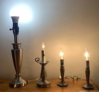 Front view of 4 silver-tone lamps with light bulbs turned on, showing varying base styles and heights.