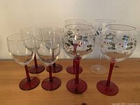 Lot of 8 wine glasses arranged showing 4 smaller plain glasses with red stems and bases and 4 larger glasses with red stems and bases and Christmas holly leaf and berry design