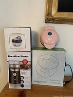 Boxed Hamilton Beach Custom Grind Coffee Grinder, boxed Multi-Purpose Manual Food Chopper, boxed Real Appeal Electronic Food Scale, and small pink pancake maker on top of boxes