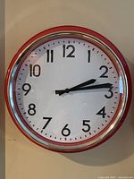 Front view of red framed round wall clock showing clock face with black numbers on white background, black hour and minute hands.